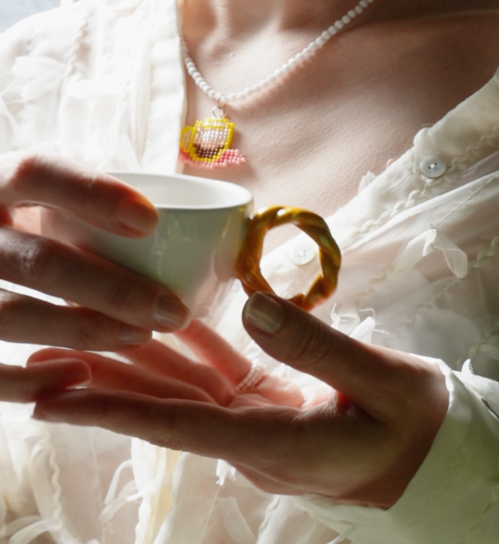 Person holding a small white cup of coffee wearing a white shirt and handmade beaded coffee necklace