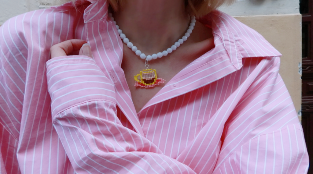 Person wearing a pink striped shirt with a white beaded necklace featuring a caffè ristretto cup pendant.