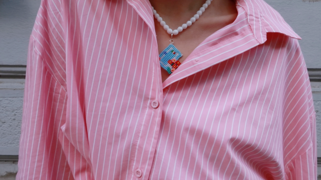 Person wearing a pink striped shirt with a white beaded necklace featuring a small Italian house pendant.