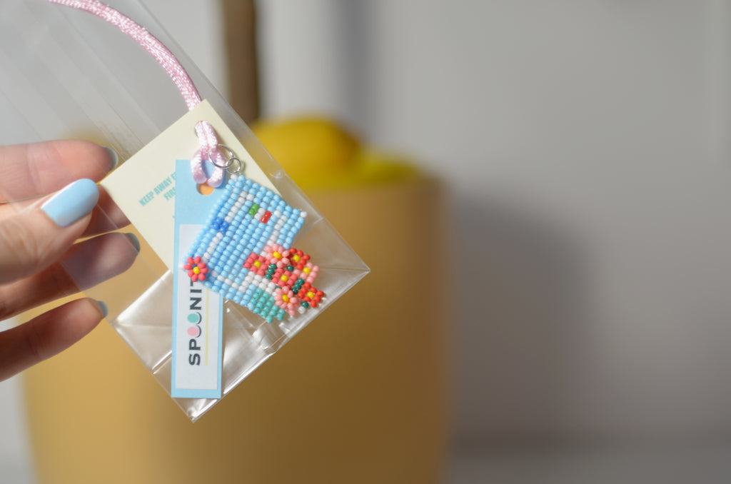 Hand holding a clear bag with a beaded Italian house pendant with blue door and geraniums, SPOONITTO branding.