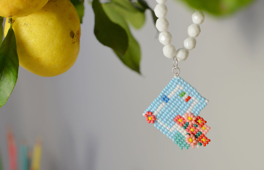 White beaded necklace with a pastel Italian house pendant next to lemons and leaves.
