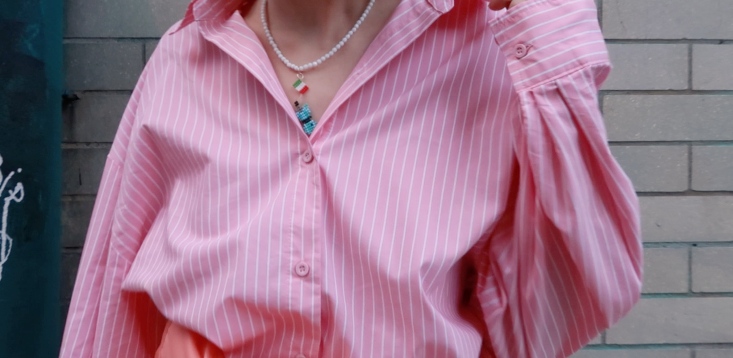 Person wearing a pink striped shirt with a beaded moka pot pendant on a blurred brick background