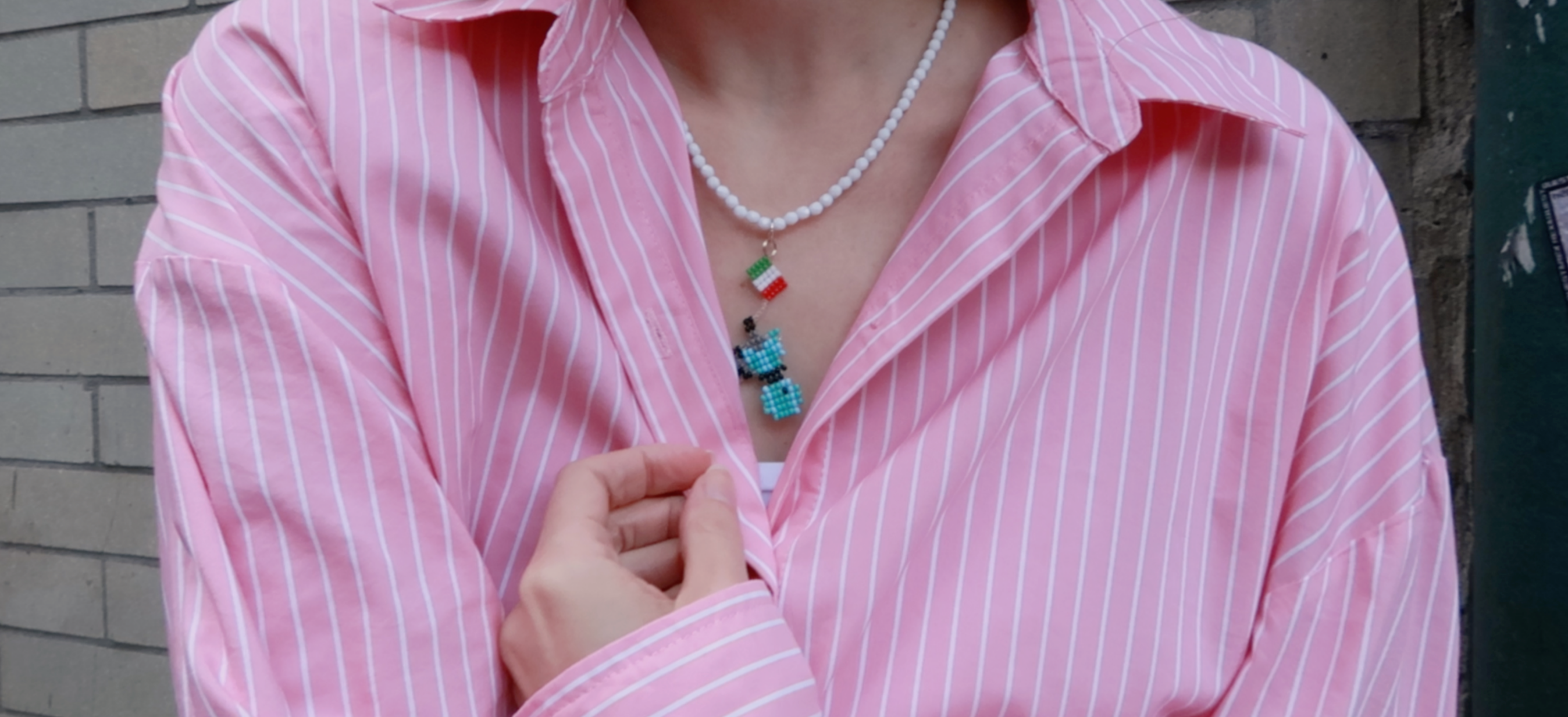 Person wearing a pink striped shirt with a pearl necklace and colorful moka pot pendant with italian flag
