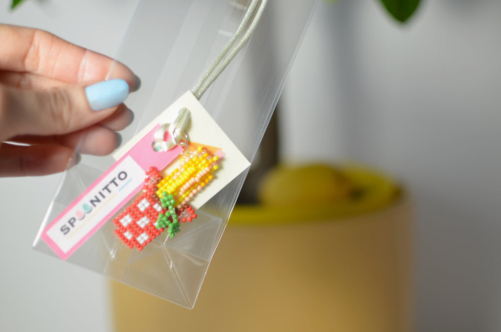 Hand holding a clear bag with a beaded spaghetti and basil pendant and SPOONITTO branding.