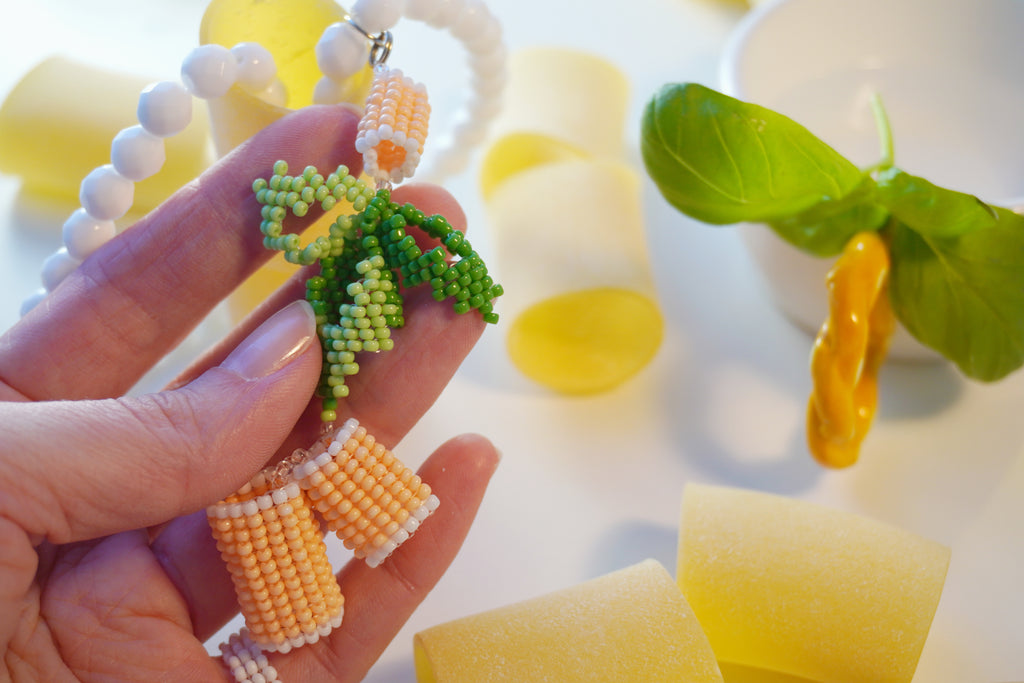 Hand holding a beaded italian food inspired necklace with green basil leaves and orange paccheri pasta, surrounded by yellow and white objects.