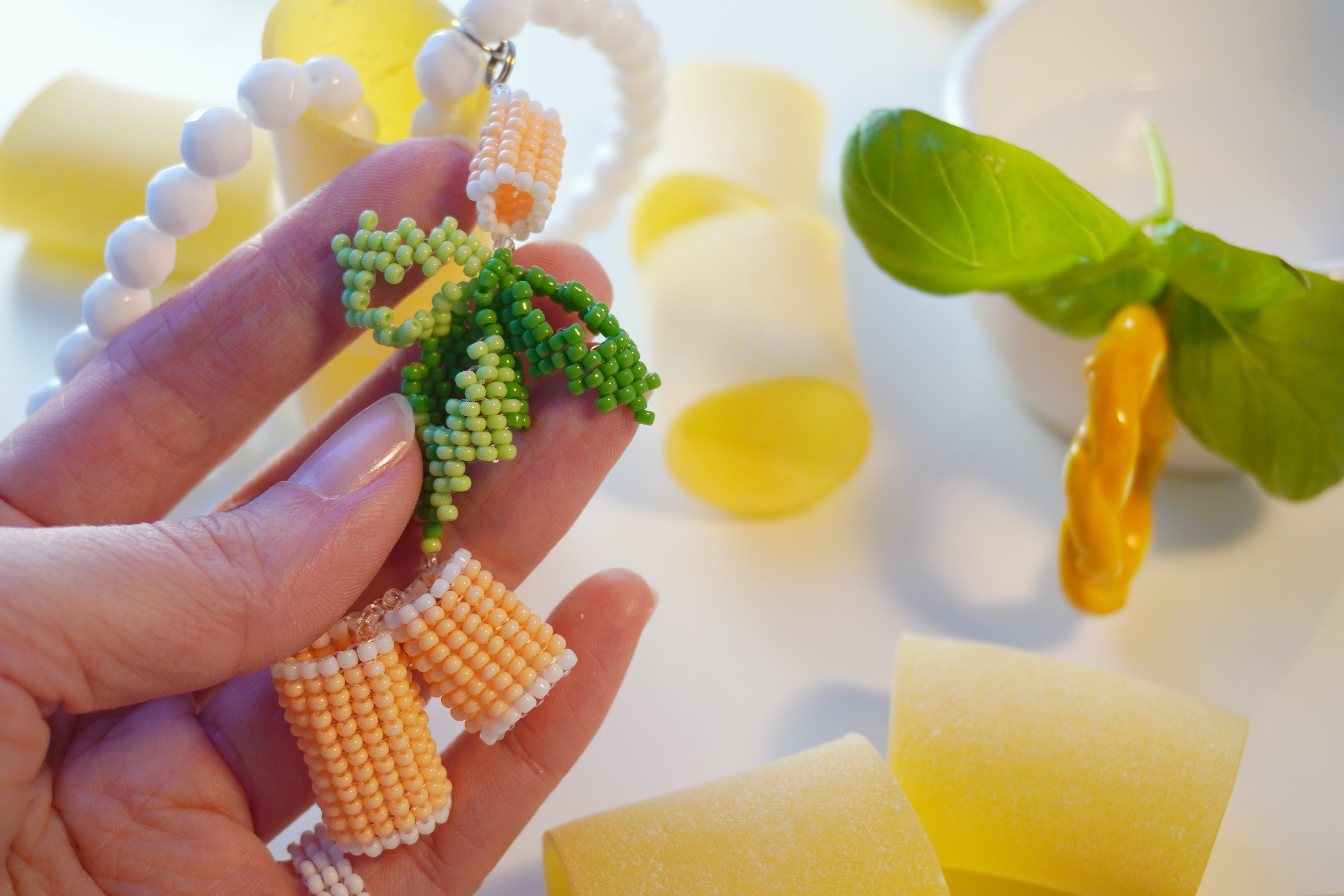 Hand holding a beaded italian food inspired necklace with green basil leaves and orange paccheri pasta, surrounded by yellow and white objects.