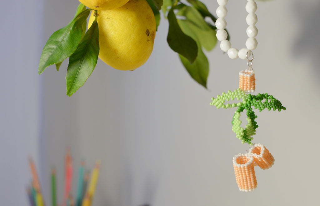 White beaded chain with a pasta and basil design pendant near lemons and leaves on a blurred background.
