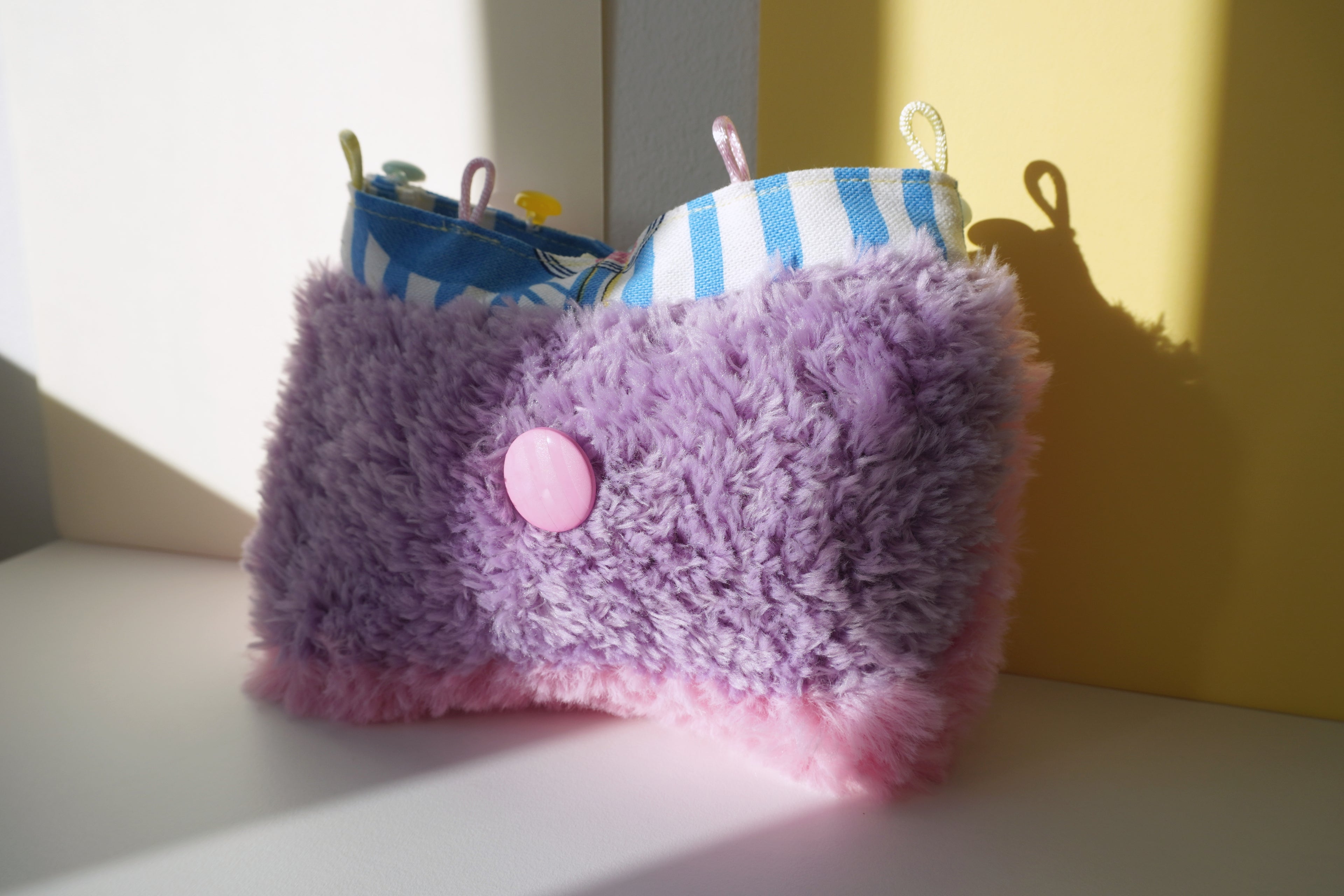 Purple fluffy purse with a pink button on a white surface with a yellow wall in the background