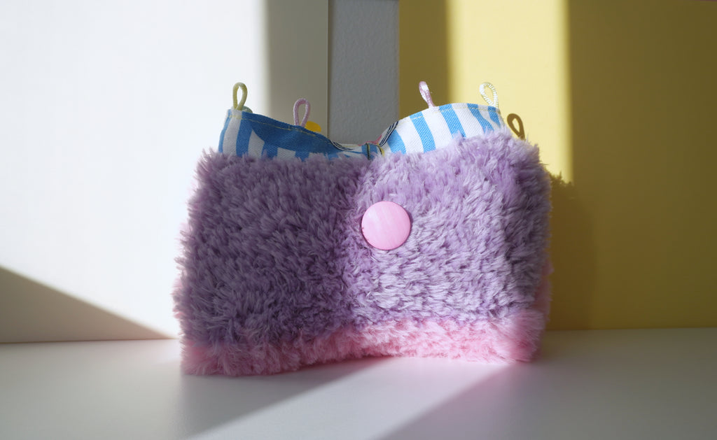 Purple and pink fluffy small bag with a button on a white surface with a yellow wall background