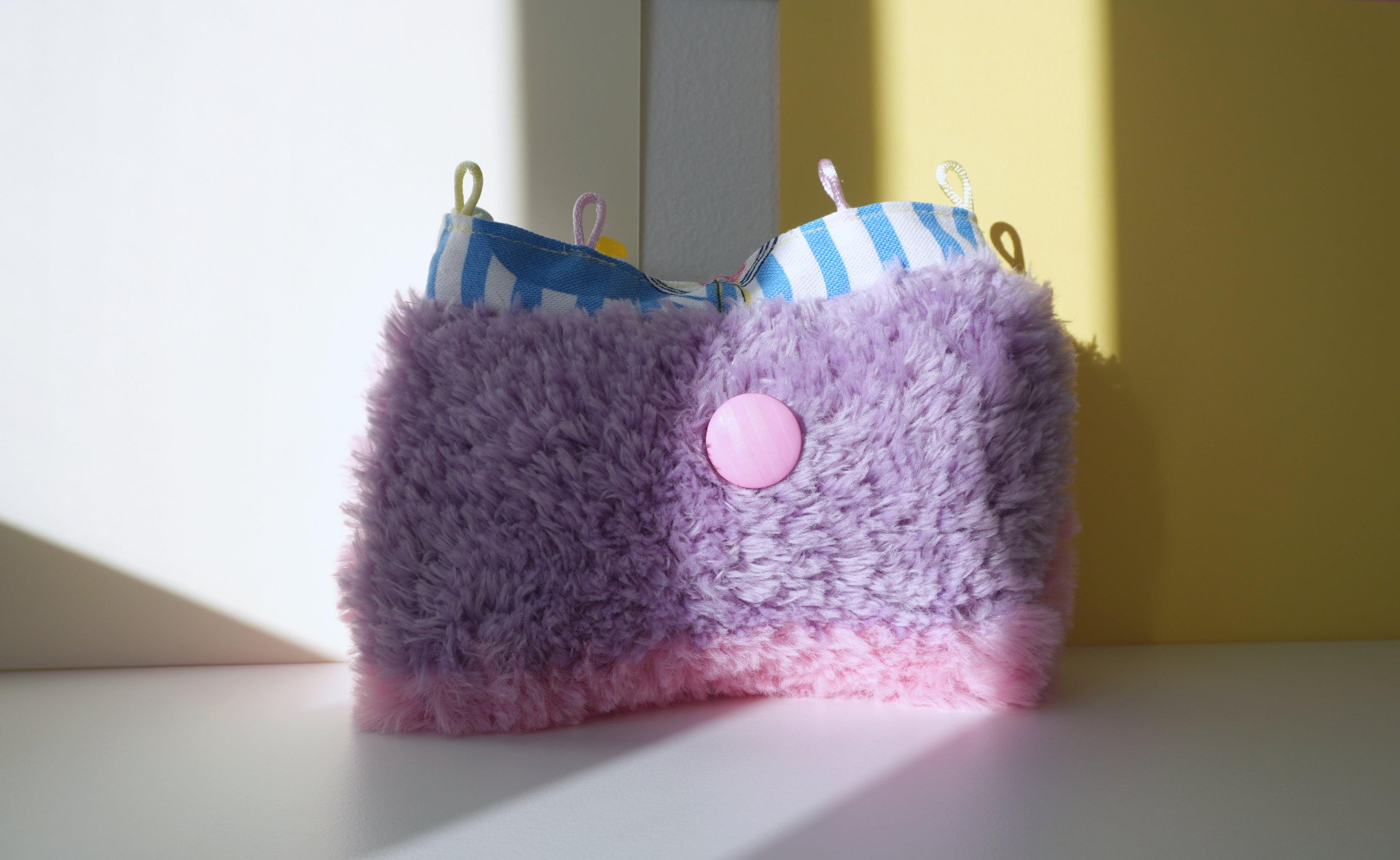 Purple and pink fluffy small bag with a button on a white surface with a yellow wall background