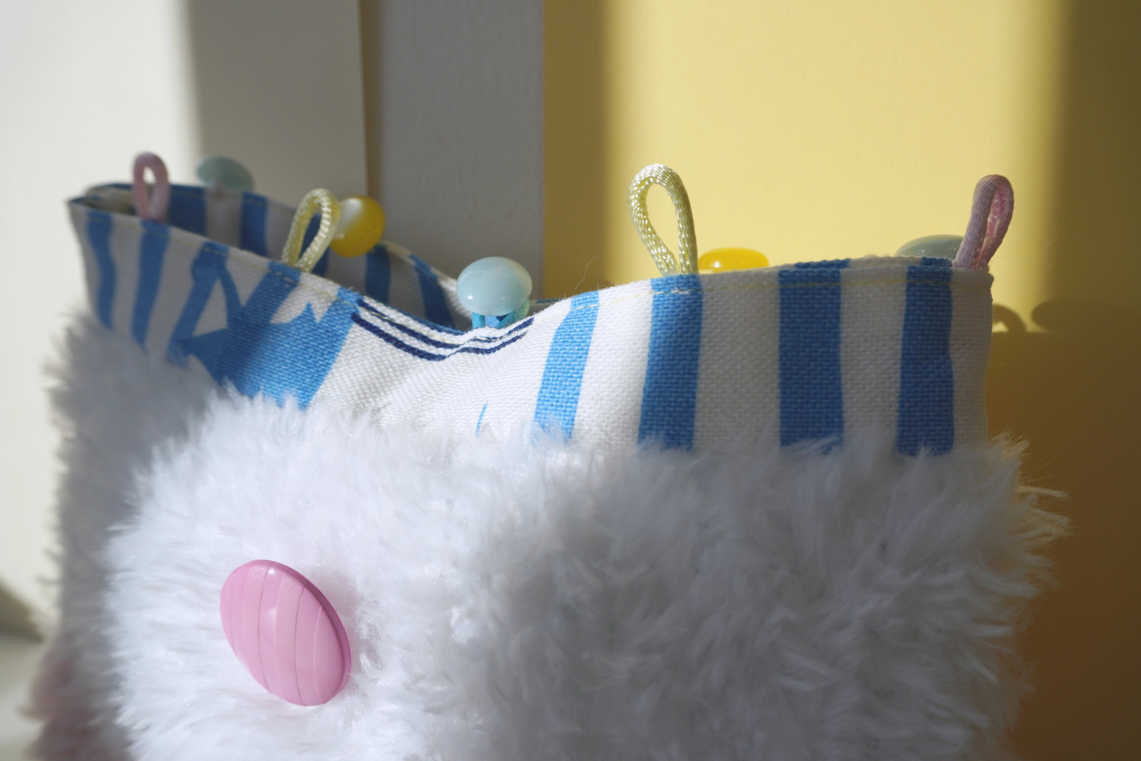 White fluffy hand-knitted small purse  with blue and white striped linen and a pink button, against a yellow background.