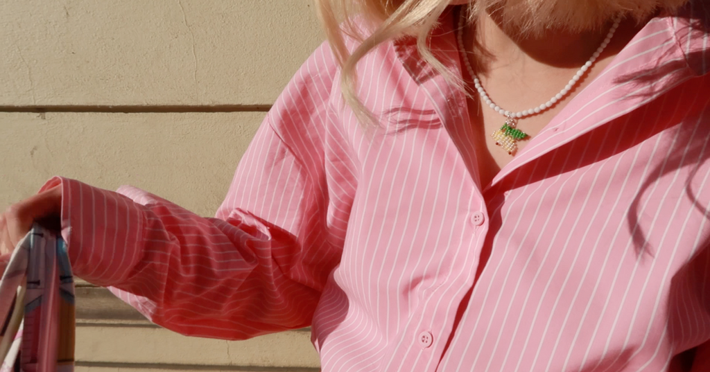 Person wearing a pink striped shirt with a beaded white necklace with lemon pendant, standing against an olive wall.
