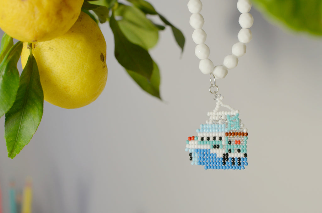 Beaded necklace with a small blue and white ship pendant next to lemons and leaves.