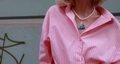Person wearing a pink striped shirt with a necklace featuring a blue ship pendant against a light green wall.
